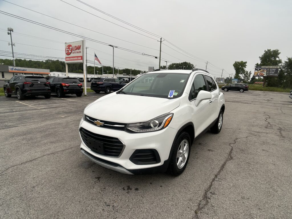 White Chevrolet SUV in the foreground at a car dealership lot, with other vehicles and an EZ LOAN sign in the background
