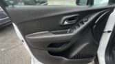 Interior of a car's front driver-side door showing the window control panel, chrome door handle, and armrest; raindrops on the window with a parking lot outside.