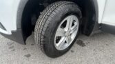 Close-up of a vehicle wheel: tire with pronounced tread, silver alloy rim, and surrounding wheel well on a light-colored fender.