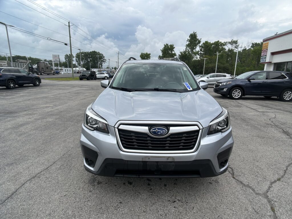 Front view of a silver Subaru SUV parked in a lot with other vehicles and a dealership building in the background under a cloudy sky.