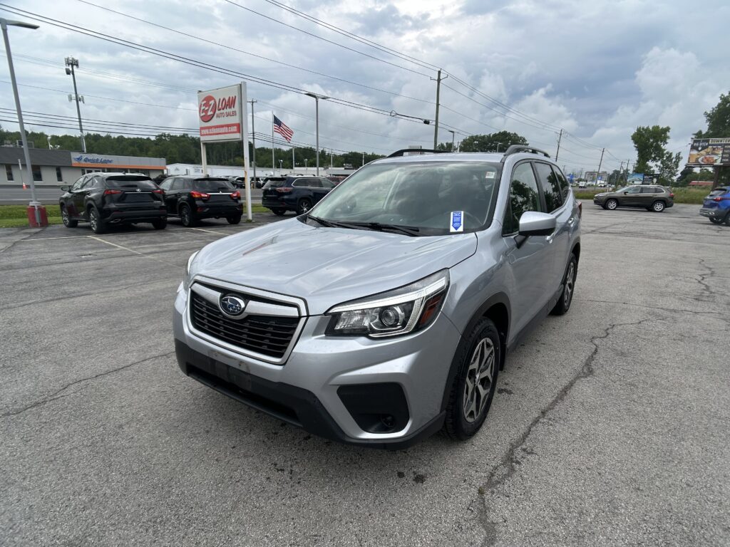 Silver SUV in the foreground at a car dealership lot with other vehicles and signs in the background under a cloudy sky.