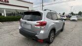 Silver Subaru Forester parked in a dealership lot beside a glass-front building with 'SALES' sign visible and multiple cars in the background.