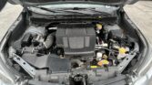 Open car hood revealing engine bay with Subaru Boxer engine, yellow oil cap, battery, and various hoses and components visible under the hood.