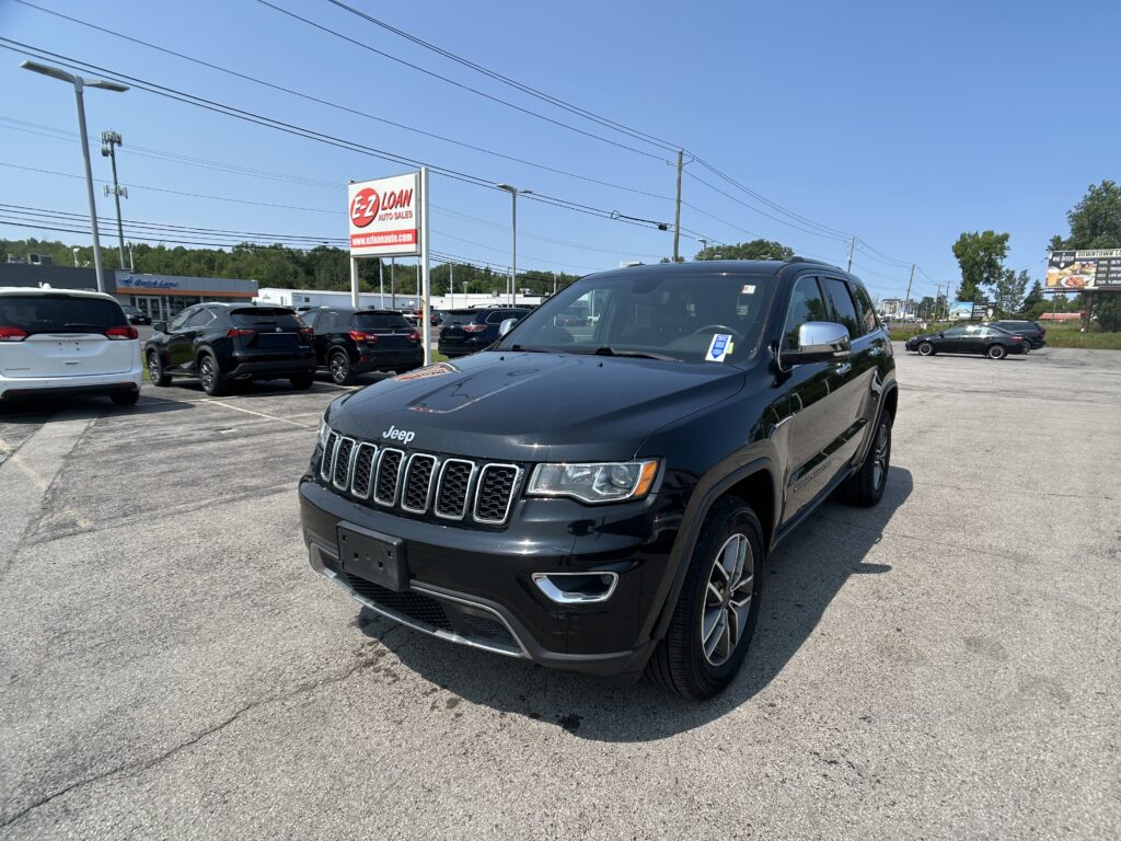 Close-up of a dark gray Jeep Grand Cherokee in a car dealership lot, with other vehicles and a bright blue sky in the background.