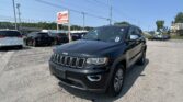Close-up of a dark gray Jeep Grand Cherokee in a car dealership lot, with other vehicles and a bright blue sky in the background.