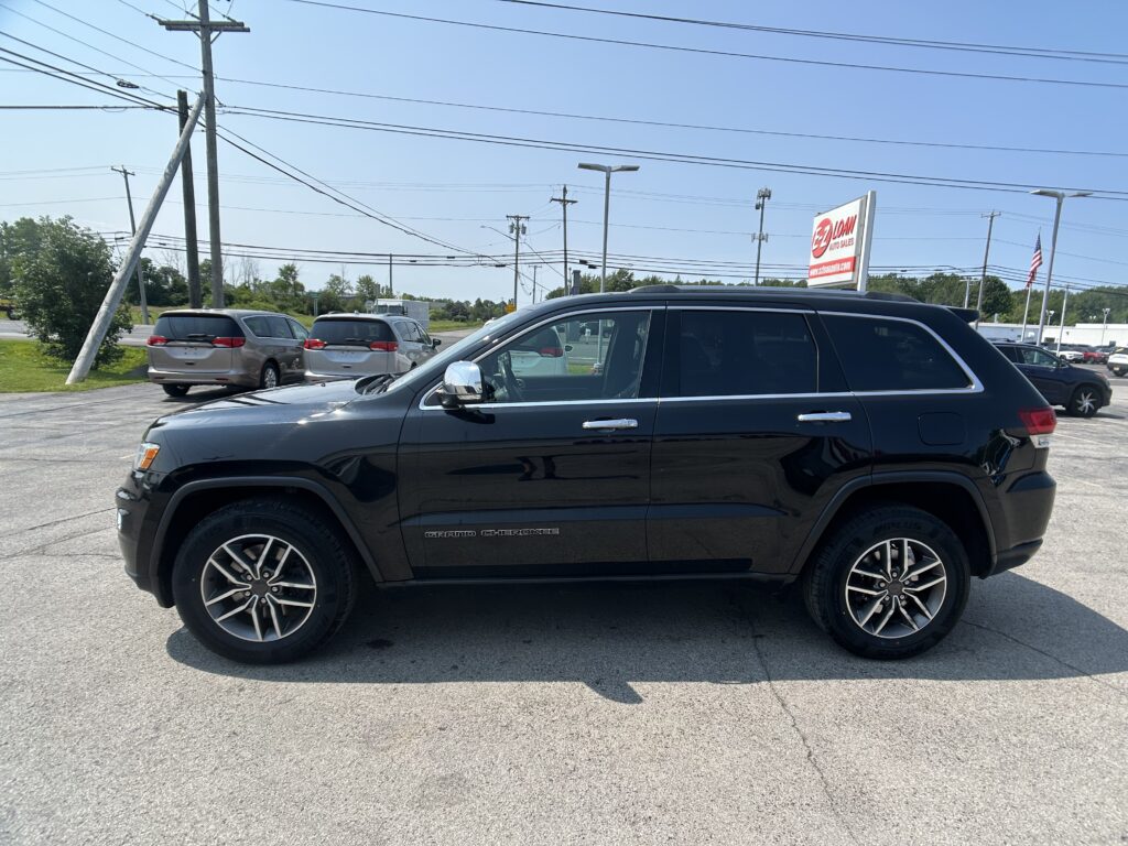 Black Jeep Grand Cherokee SUV parked in a car lot with other vehicles and a showing sign in background reading 'Auto Sales'