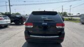 Rear view of a black Jeep SUV in a car dealership lot with power lines overhead and other vehicles nearby.