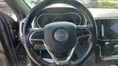 Driver's-seat view of a Jeep steering wheel with gauge cluster and infotainment screen, overlooking a car lot through the windshield.