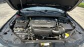 Car engine bay with hood open, showing battery, air intake, and various hoses visible under the open hood.