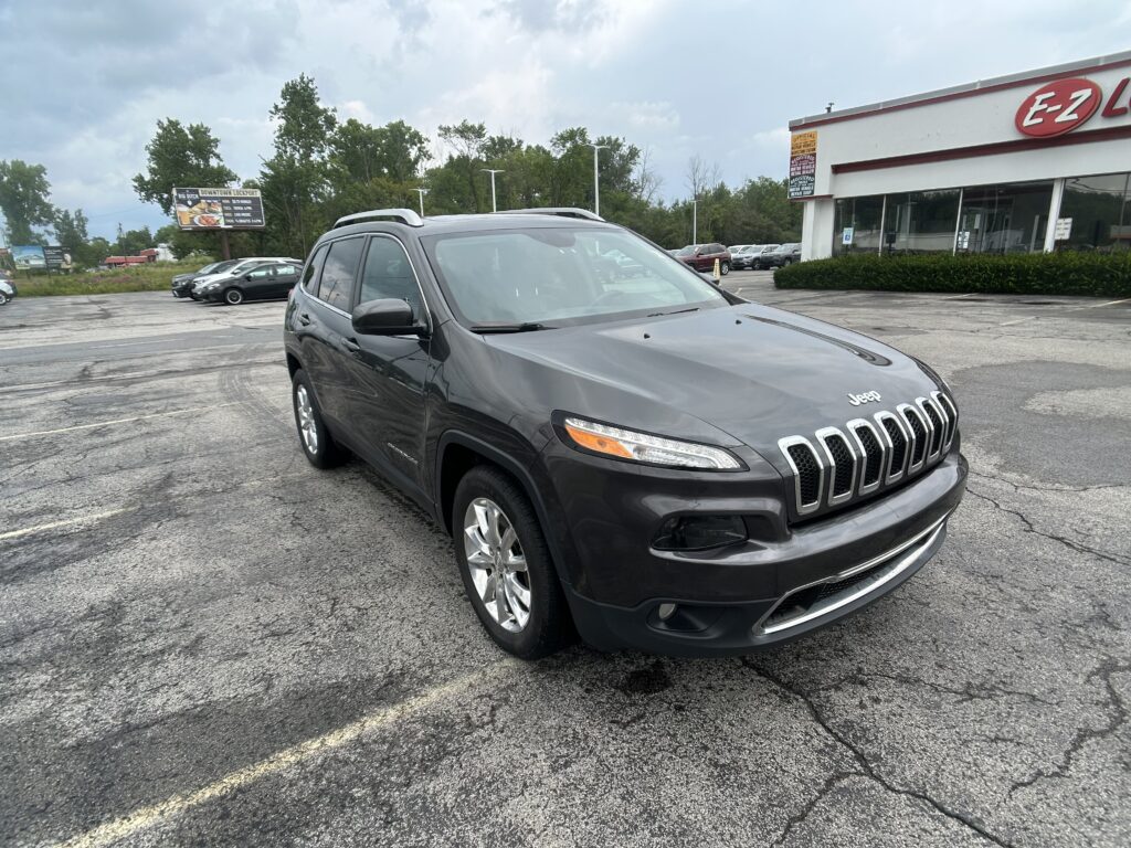 Dark gray Jeep SUV parked in a lot near an EZ Lube service building with other cars and trees in the background, cloudy sky above.