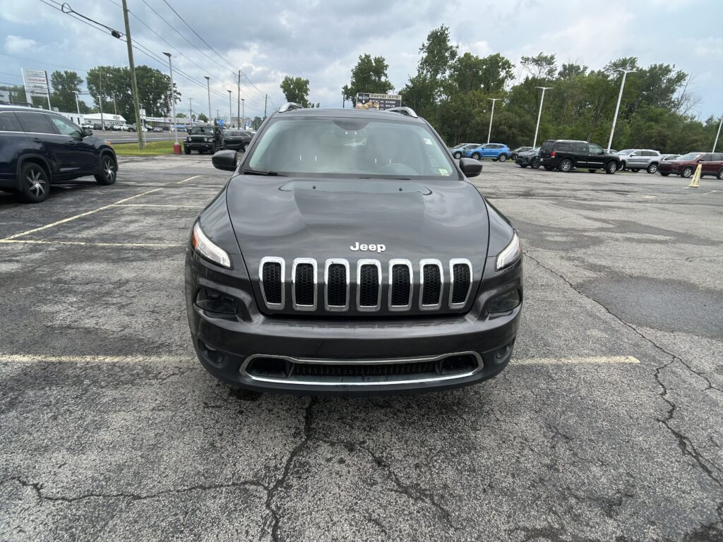 Front view of a gray Jeep SUV parked in a crowded lot, with other cars and trees in the background.