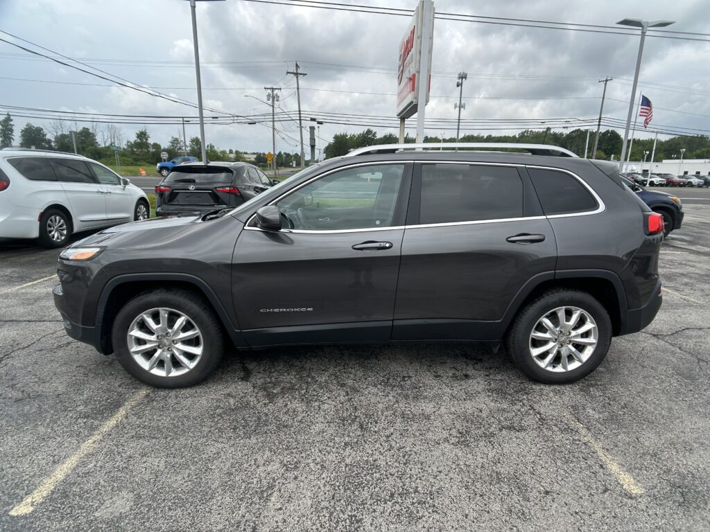 Side view of a dark gray Jeep Cherokee SUV parked in a busy car lot with other vehicles behind it and a cloudy sky above.