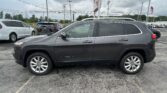 Side view of a dark gray Jeep Cherokee SUV parked in a busy car lot with other vehicles behind it and a cloudy sky above.