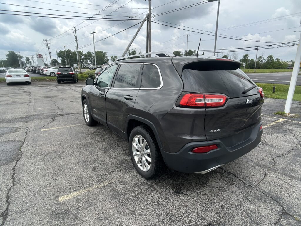 Rear three-quarter view of a gray Jeep SUV with roof rails in a parking lot, power lines overhead and a cloudy sky.