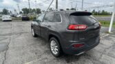 Rear three-quarter view of a gray Jeep SUV with roof rails in a parking lot, power lines overhead and a cloudy sky.
