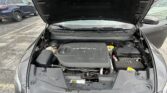 Open car hood showing engine bay in a parking lot, with other vehicles nearby and windshield wipers visible on the windshield