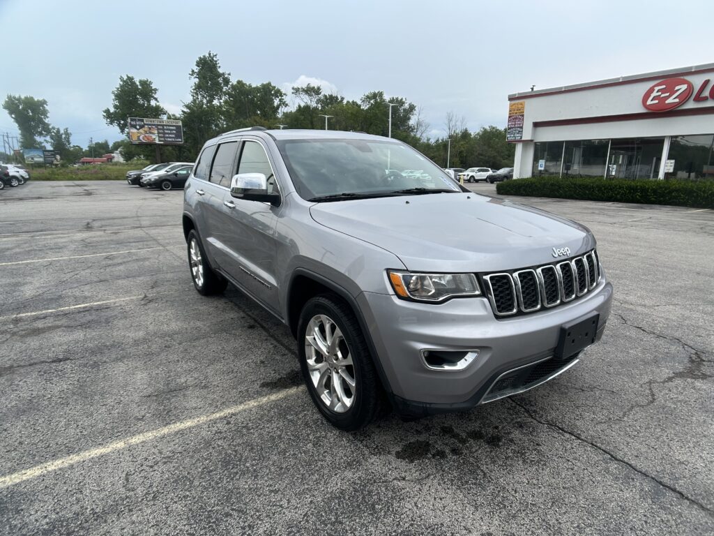 Front-right view of a silver Jeep SUV parked in a dealership lot, with a white building and red E-Z Auto sign in the background.