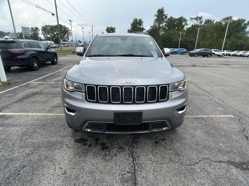 Front view of a silver Jeep SUV parked in a dealership lot, with other cars and trees in the background.