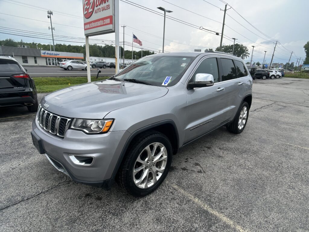 Silver Jeep Grand Cherokee in a car dealership lot with other vehicles and a tall red-and-white sign near the foreground, American flag waving in the background.