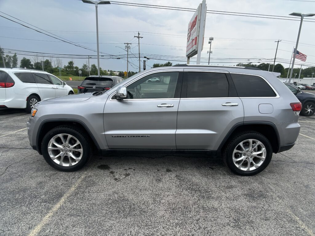 Side view of a silver Jeep Grand Cherokee parked in a lot with other vehicles and overhead power lines in the background.