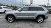 Side view of a silver Jeep Grand Cherokee parked in a lot with other vehicles and overhead power lines in the background.