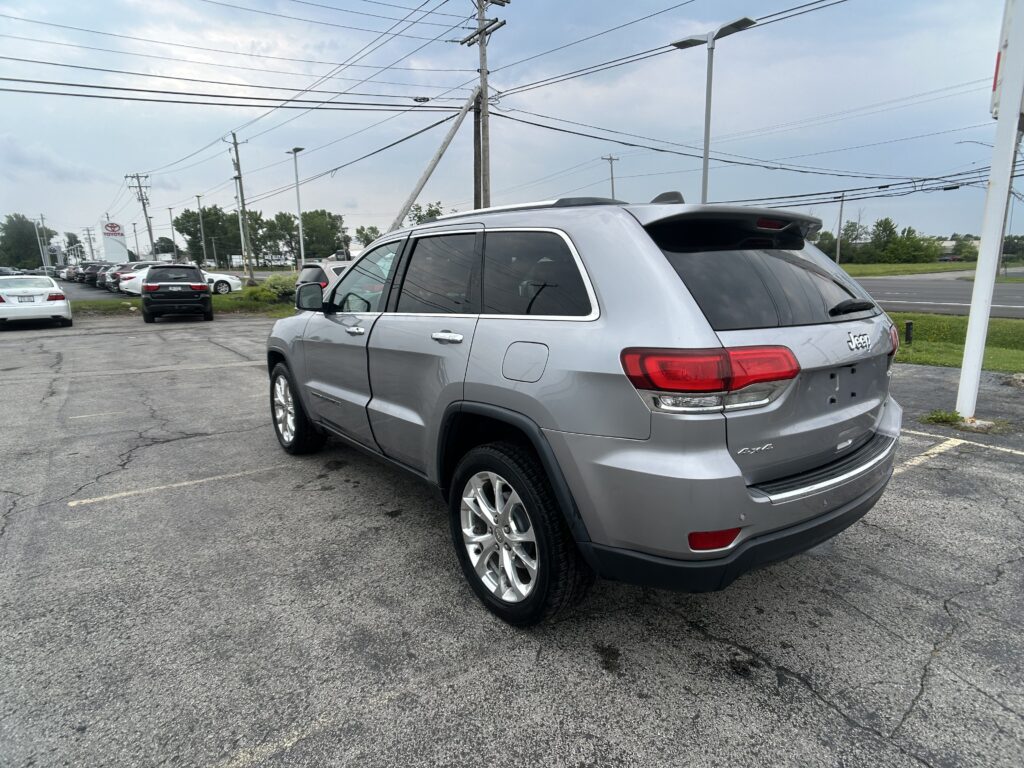 Rear three-quarter view of a gray Jeep SUV parked in a car dealership lot with power lines overhead and other cars in the background.
