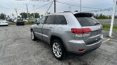 Rear three-quarter view of a gray Jeep SUV parked in a car dealership lot with power lines overhead and other cars in the background.