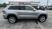 Silver Jeep Grand Cherokee SUV parked in a dealership lot with an 'E-Z LOAN AUTO SALES' sign on the building in the background, other cars visible behind.