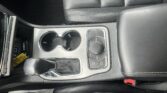 Center console in a car with two cup holders, gear selector, and control dial surrounded by black leather seats.