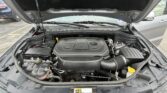 Open car hood revealing engine bay with engine cover in the center, battery on the left, and various hoses and wiring visible under the dashboard edge.
