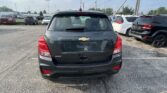 Rear view of a gray Chevrolet Trax SUV in a car dealership lot with other vehicles lining the row behind it and power lines overhead.