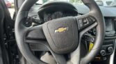 Close-up of a black perforated steering wheel with Chevrolet emblem; the dashboard and infotainment screen are visible behind it. Outside cars can be seen through the windshield and open door on the left.