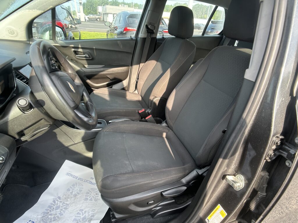 Front interior of a car showing the steering wheel on the left, two black fabric seats, a center console, and a paper floor mat on the driver side; outside view of other cars through the windows.