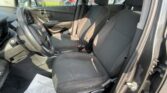 Front interior of a car showing the steering wheel on the left, two black fabric seats, a center console, and a paper floor mat on the driver side; outside view of other cars through the windows.