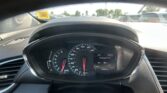 View from the driver's seat: steering wheel and instrument cluster with speedometer, tachometer, and digital display; outside are parked cars and a yellow windshield sticker.