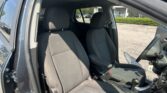 Open-side view of a car’s front seats and driver-side door, showing black fabric upholstery and a sunny outdoor lot beyond the window.