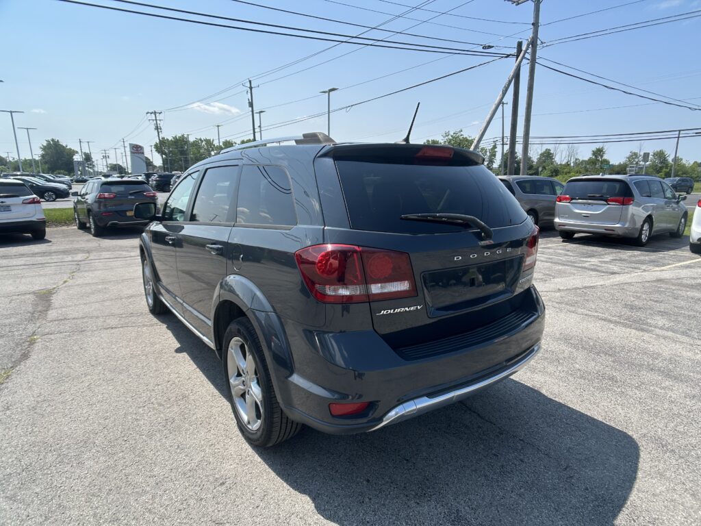 Rear three-quarter view of a dark gray Dodge Journey SUV in a busy parking lot on a sunny day, with other cars and power lines in the background.