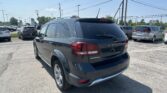Rear three-quarter view of a dark gray Dodge Journey SUV in a busy parking lot on a sunny day, with other cars and power lines in the background.