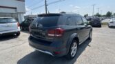 Dark gray Dodge Durango Crossroad SUV in a dealership parking lot, with a white building on the left and a row of cars under a blue sky.