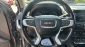 Driver's-seat view of a GMC vehicle: steering wheel centered, dashboard gauges visible, center console touchscreen to the right, passenger door open, sunny parking lot outside with other cars.