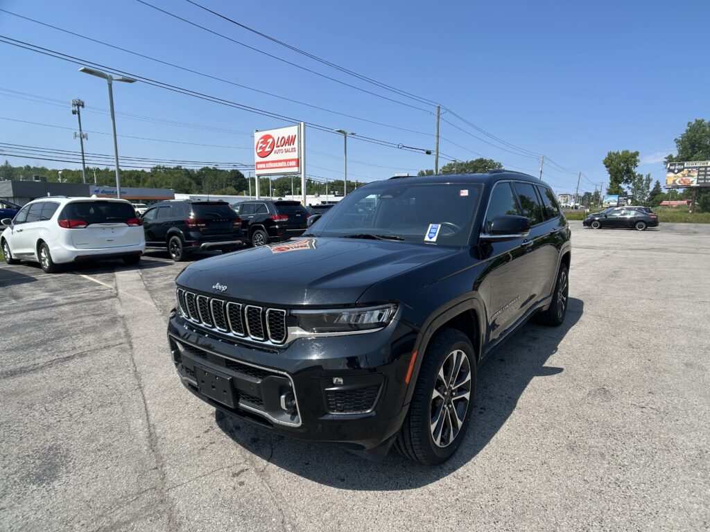 Black Jeep Grand Cherokee in a dealership lot with other cars and an EZ Loan Auto Sales sign against a clear blue sky.