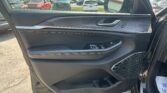 Close-up of a car's interior door panel featuring a chrome handle, wood-grain trim, and window/mirror controls; parking lot outside visible through the window.