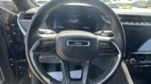 Close-up of Jeep steering wheel with white stitching, digital dashboard visible behind it and car controls on both sides, outdoors through the windshield.