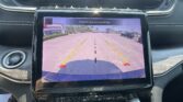 In-car infotainment screen showing a rearview camera view with parking lot, yellow/green guiding lines, and the warning text 'Check Entire Surroundings'