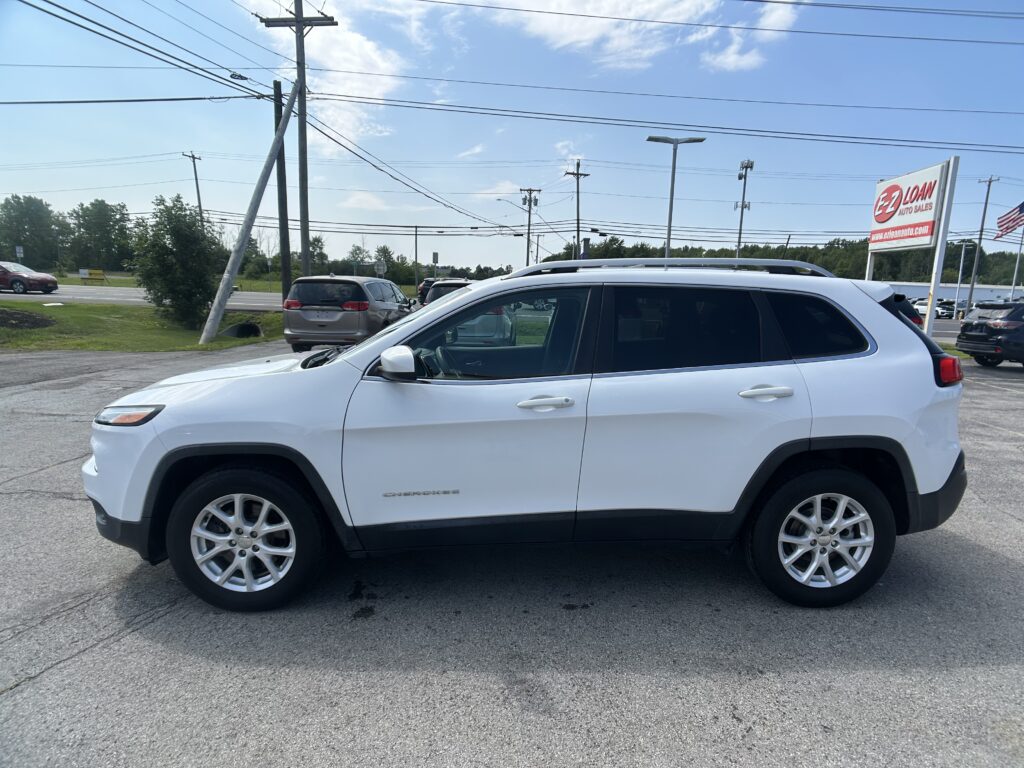 White SUV (Jeep Grand Cherokee) parked in a dealership lot, with an E-LOAN Auto Sales sign and American flag in the background on a sunny day.