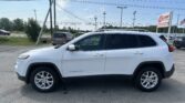 White SUV (Jeep Grand Cherokee) parked in a dealership lot, with an E-LOAN Auto Sales sign and American flag in the background on a sunny day.