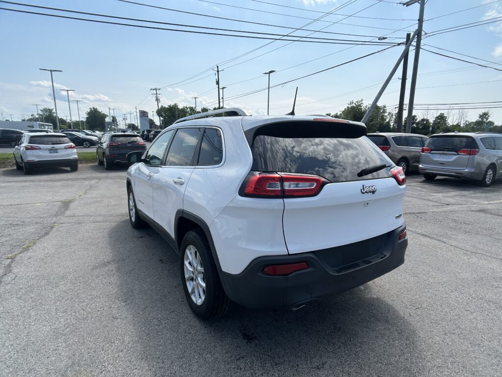 White Jeep SUV in the foreground at a car dealership lot with other vehicles and power lines overhead, under a clear sky.