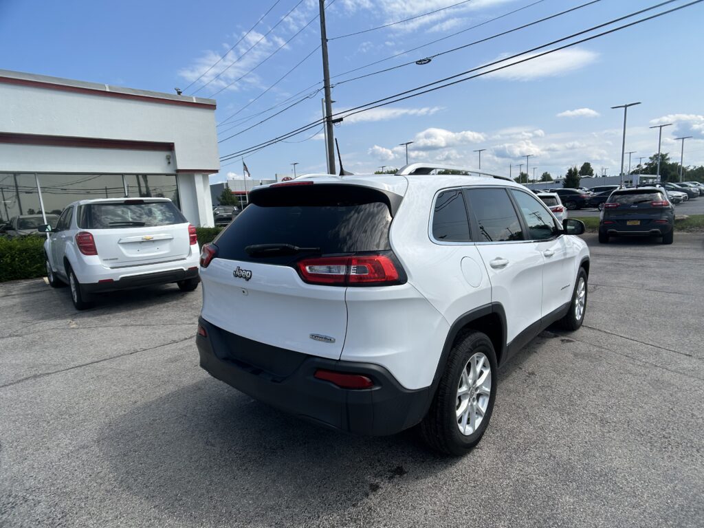 White Jeep SUV in a dealership lot, with a white building on the left and a row of parked cars under a blue sky.
