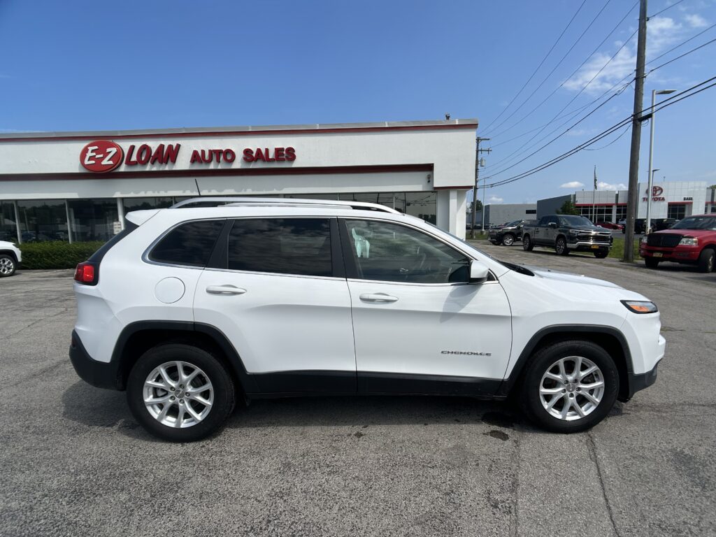 White SUV (Jeep Cherokee) parked at a car dealership with an E-Z LOAN AUTO SALES sign on the building and a clear blue sky above.