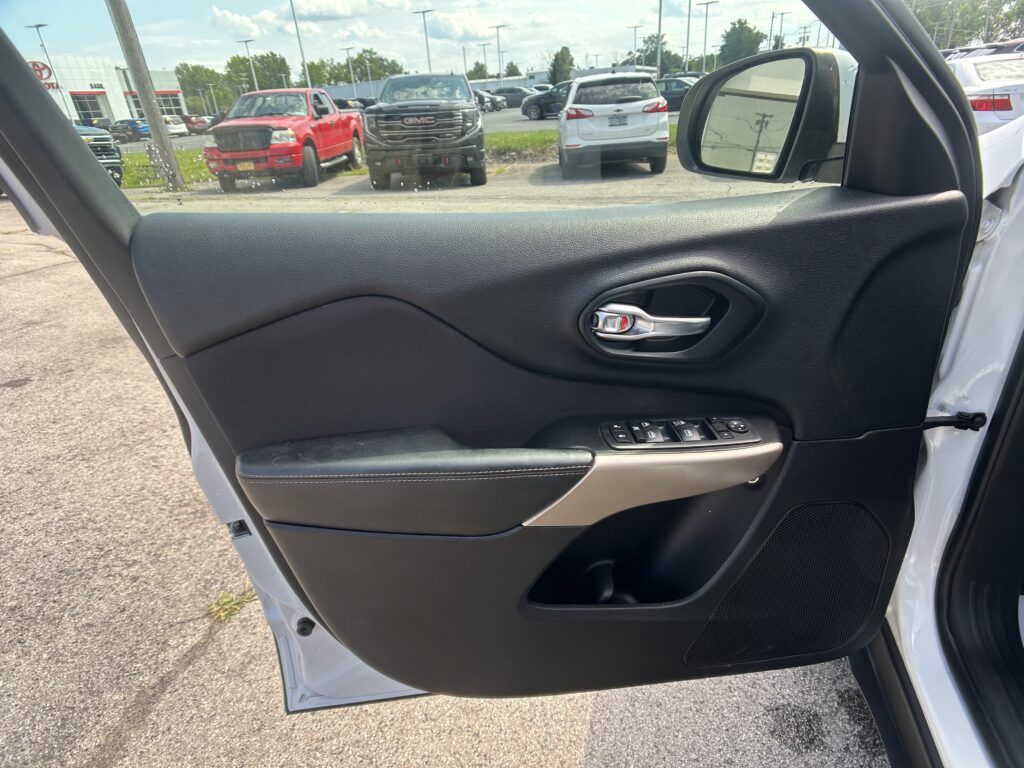 Close-up of a car door interior featuring a black textured panel, chrome door handle, and window controls; outside is a parking lot with trucks and cars visible.